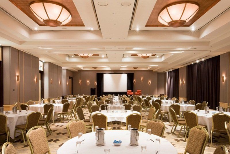 Amora Hotel Jamison Sydney Zewnętrze zdjęcie The photo shows a large banquet or conference room. There are several round tables arranged throughout the space, each covered with white tablecloths. The tables are set with water pitchers and glasses. In the center of the room, there is a projectio